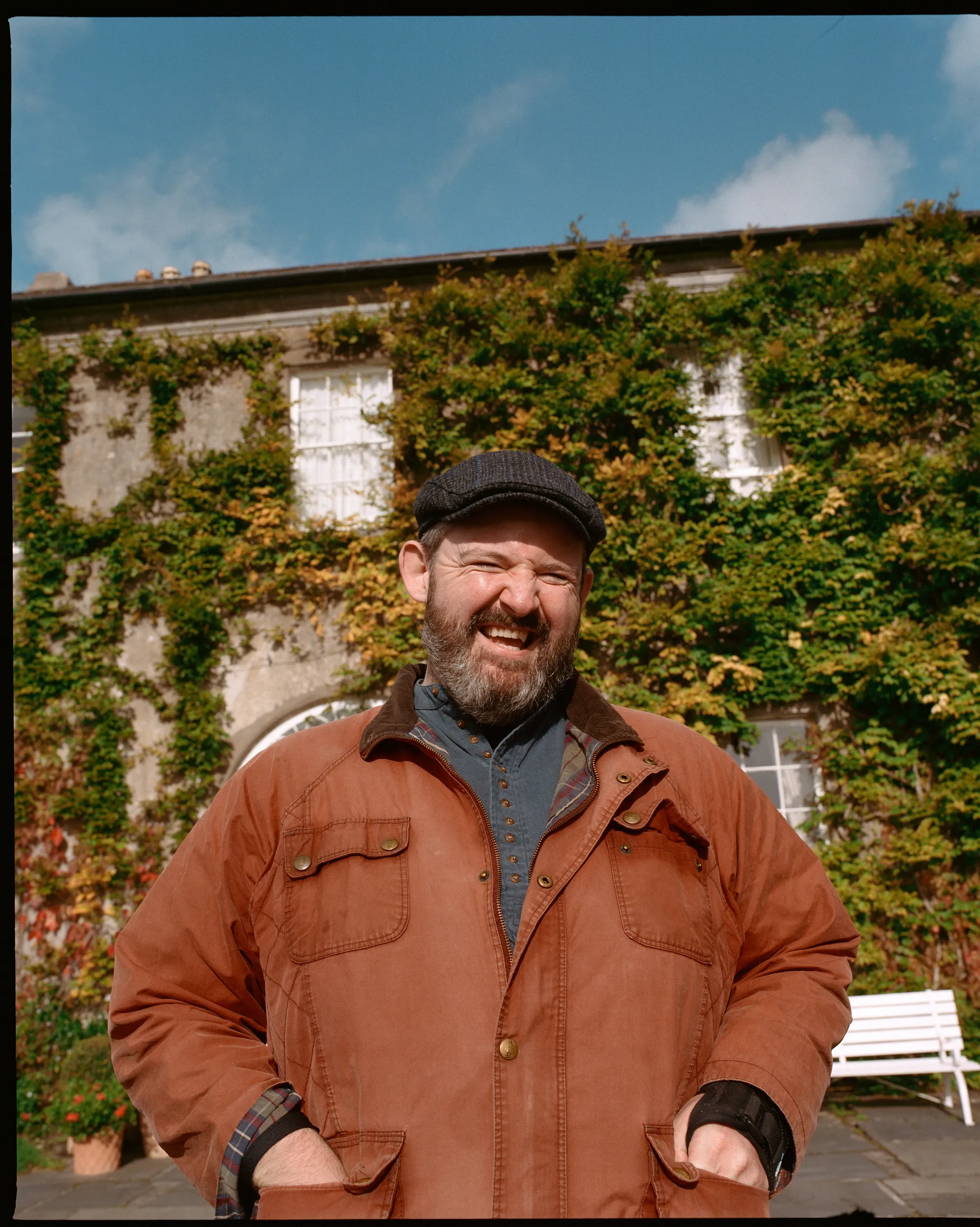 A smiling man in a flat cap outside an ivy-covered house in Ireland