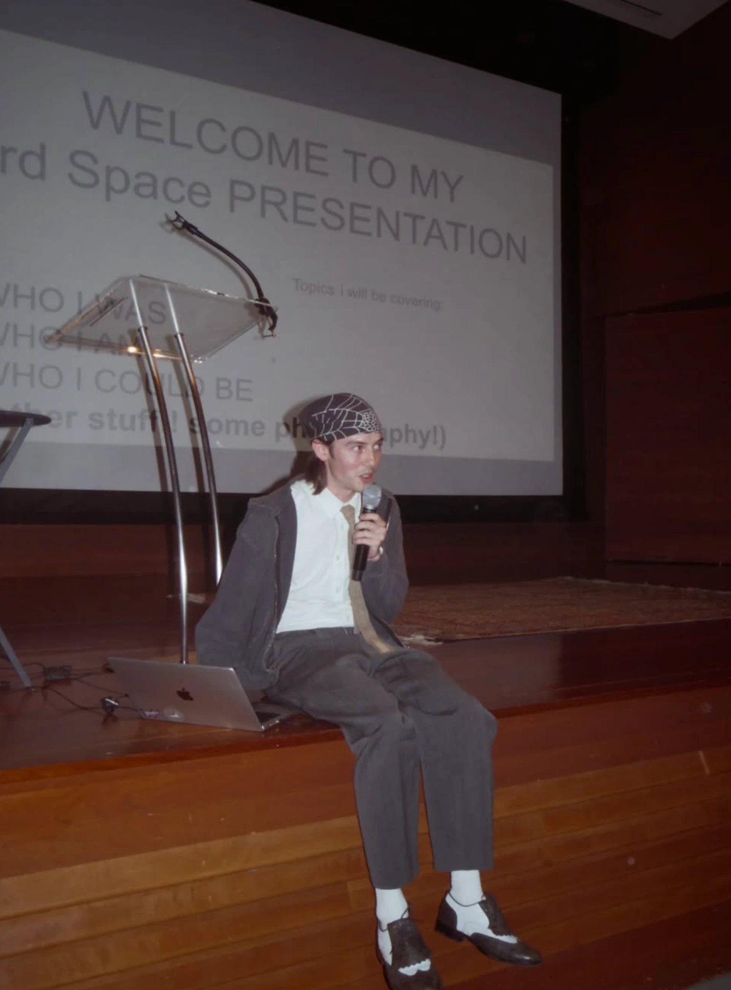 A speaker sitting on the edge of a stage beneath a slide reading “Welcome to my 3rd Space presentation”
