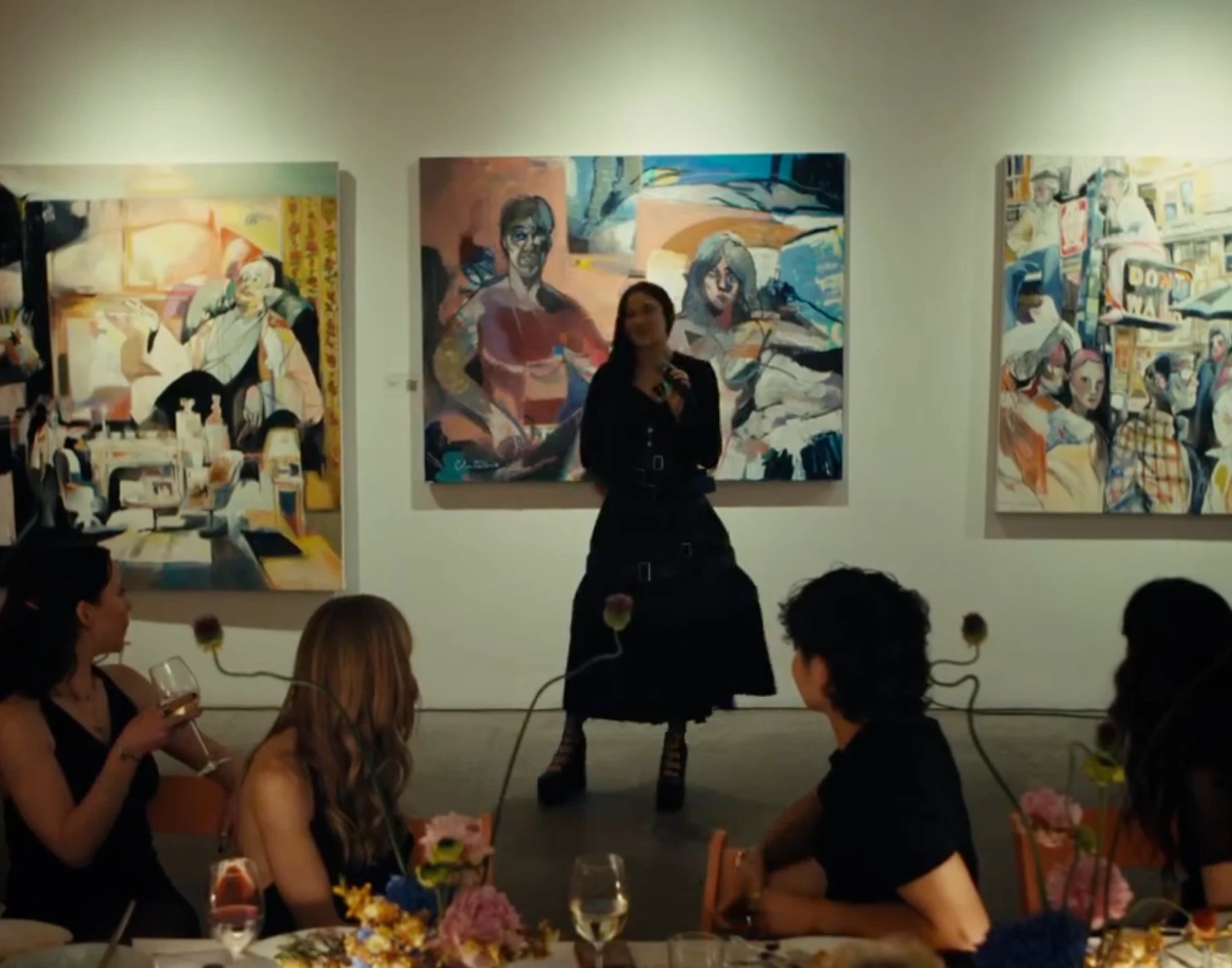 A speaker standing between paintings as guests at the table listen